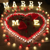 A heart-shaped display made of warm white LED alphabet letters spelling 'MARRY ME' surrounded by red and white lights.
