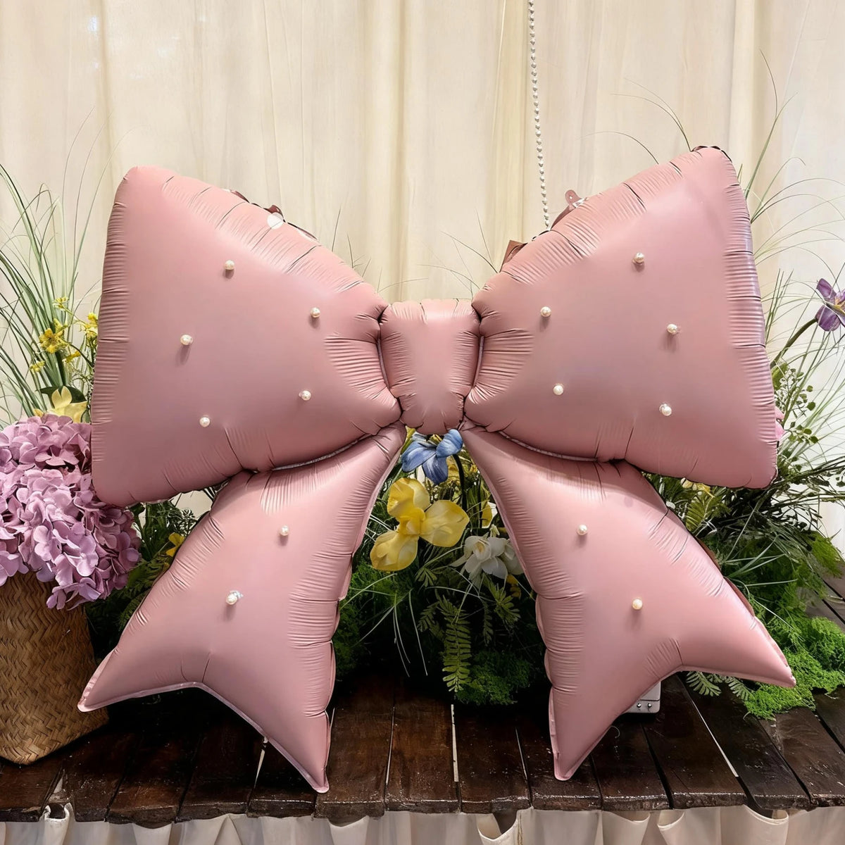 A large pink pearl bow balloon weight is displayed against a backdrop of greenery and flowers.