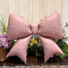 A large pink pearl bow balloon weight is displayed against a backdrop of greenery and flowers.