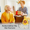 A young girl smiles while holding a gift box, with an older woman looking at the gift. The plaque is shown in the corner.