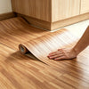 A hand is laying a roll of realistic wood grain floor stickers onto a light wooden floor.