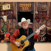 A man in a cowboy hat and shirt plays an acoustic guitar in front of a rustic wooden backdrop with a saloon sign.