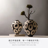 A black and cream ceramic vase with a geometric circle design sits on a wooden surface next to a dried plant branch.