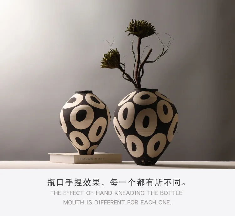 A black and cream ceramic vase with a geometric circle design sits on a wooden surface next to a dried plant branch.