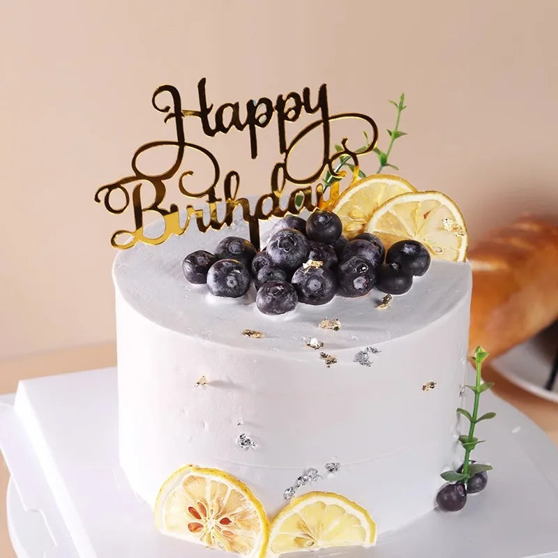 A white birthday cake decorated with fresh blueberries and a gold script topper.