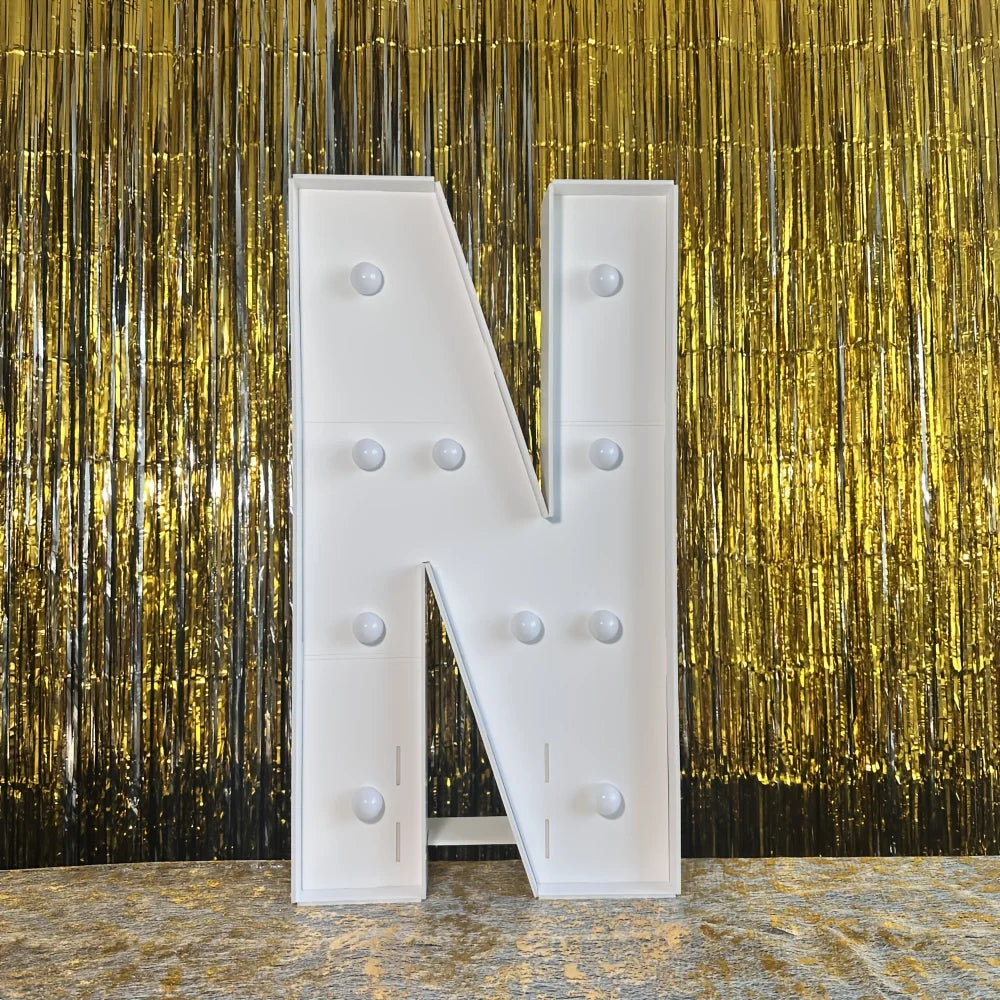 A large white marquee letter N with light-up bulbs stands against a gold metallic curtain backdrop.