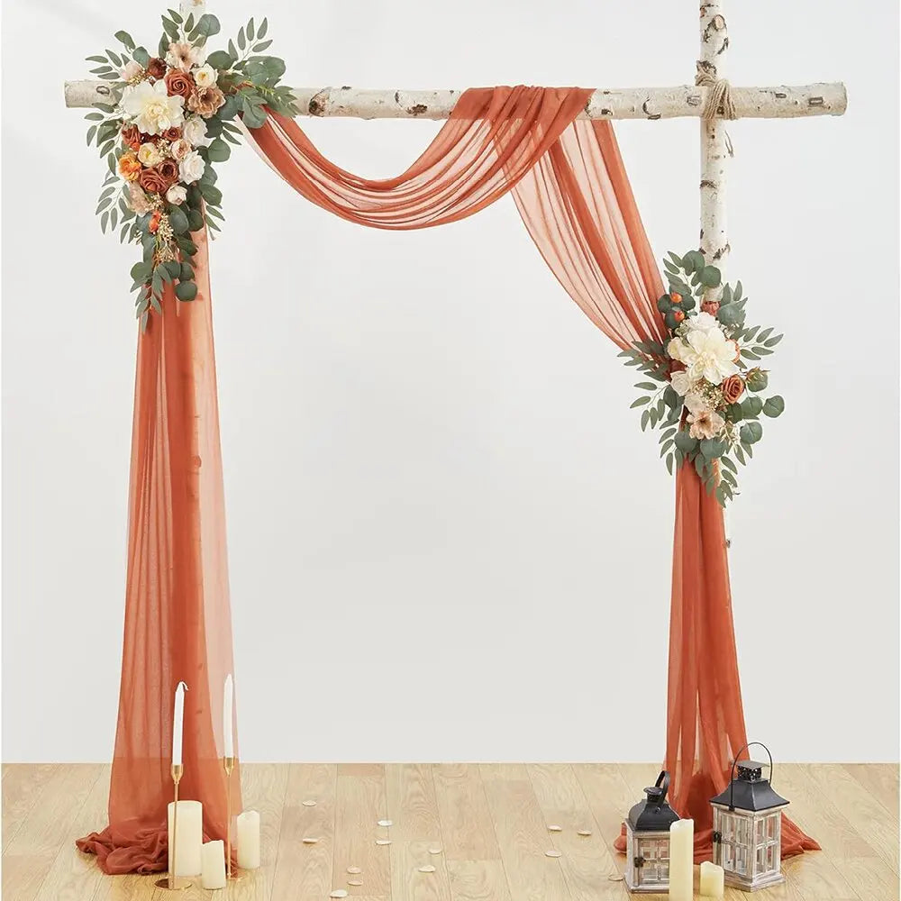 A rustic wedding arch made of branches draped with flowing burnt orange chiffon fabric and floral greenery.