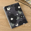 A spiral-bound notebook featuring a dark gray and black watercolor pattern with white butterflies and stars on a wooden surfa