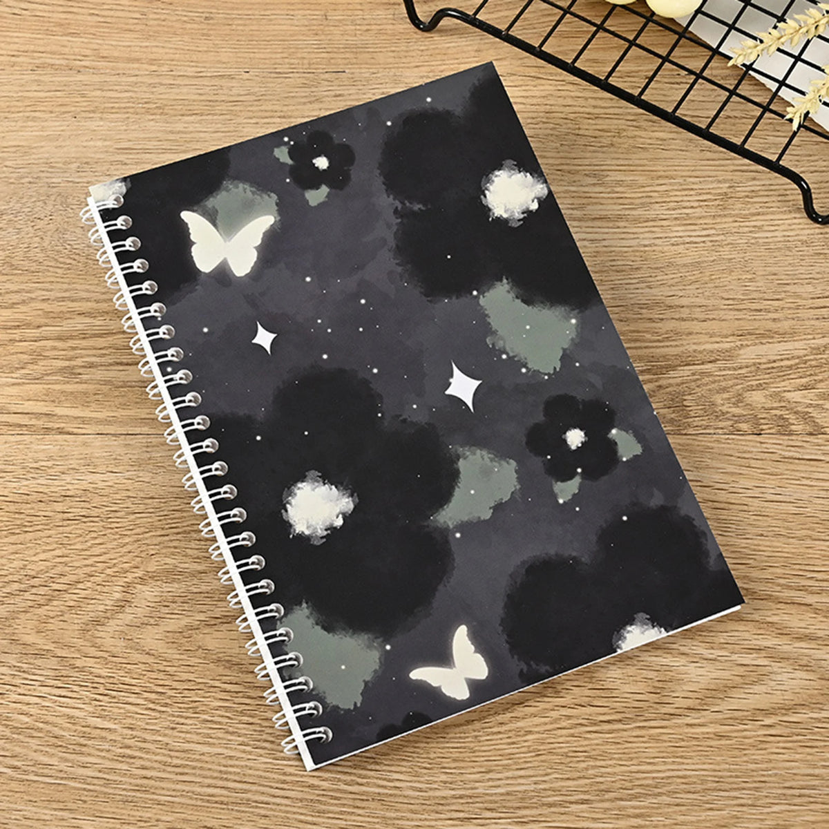 A spiral-bound notebook featuring a dark gray and black watercolor pattern with white butterflies and stars on a wooden surfa