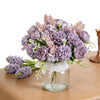 A bouquet of artificial silk peony flowers in shades of purple and lavender arranged in a clear glass vase.