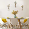 Three glass vases with dried flowers are displayed on a white wall above a wooden table.
