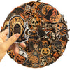 A hand holds a round sticker featuring a dark, retro graffiti aesthetic design with skulls and Halloween elements.