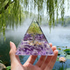 Hand holding a clear resin pyramid with purple crystals and an internal copper coil, set against a misty natural background.