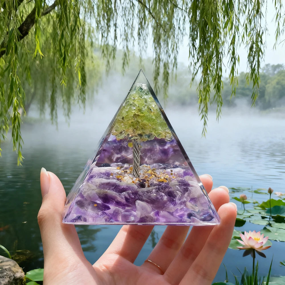 Hand holding a clear resin pyramid with purple crystals and an internal copper coil, set against a misty natural background.
