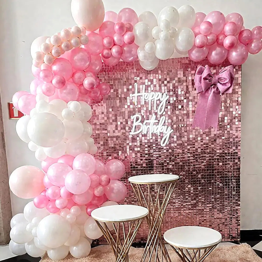 A pink and white sequin wall backdrop decorated with clusters of pink and white balloons and a pink bow for a birthday party.