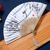 A vintage silk folding hand fan with blue and black ink drawings of bamboo on a white background resting on a wooden surface.