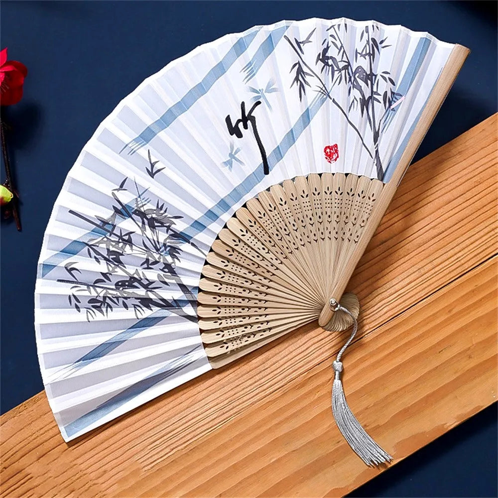 A vintage silk folding hand fan with blue and black ink drawings of bamboo on a white background resting on a wooden surface.