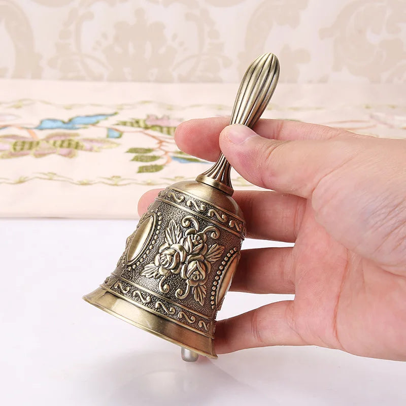 A hand is holding an ornate brass hand call bell with a decorative floral design.