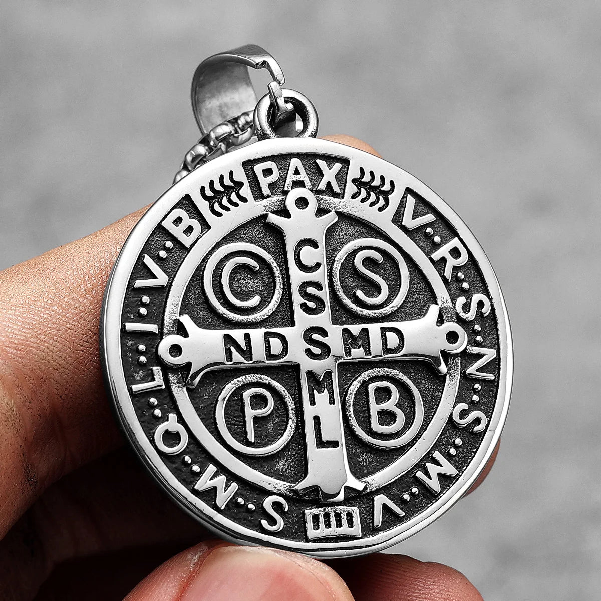 A silver stainless steel punk cross necklace pendant is held in a hand against a gray background.