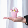 Eine Hand hält einen rosa Topf mit künstlichen rosa und weißen Blumen vor einem hellen Hintergrund.