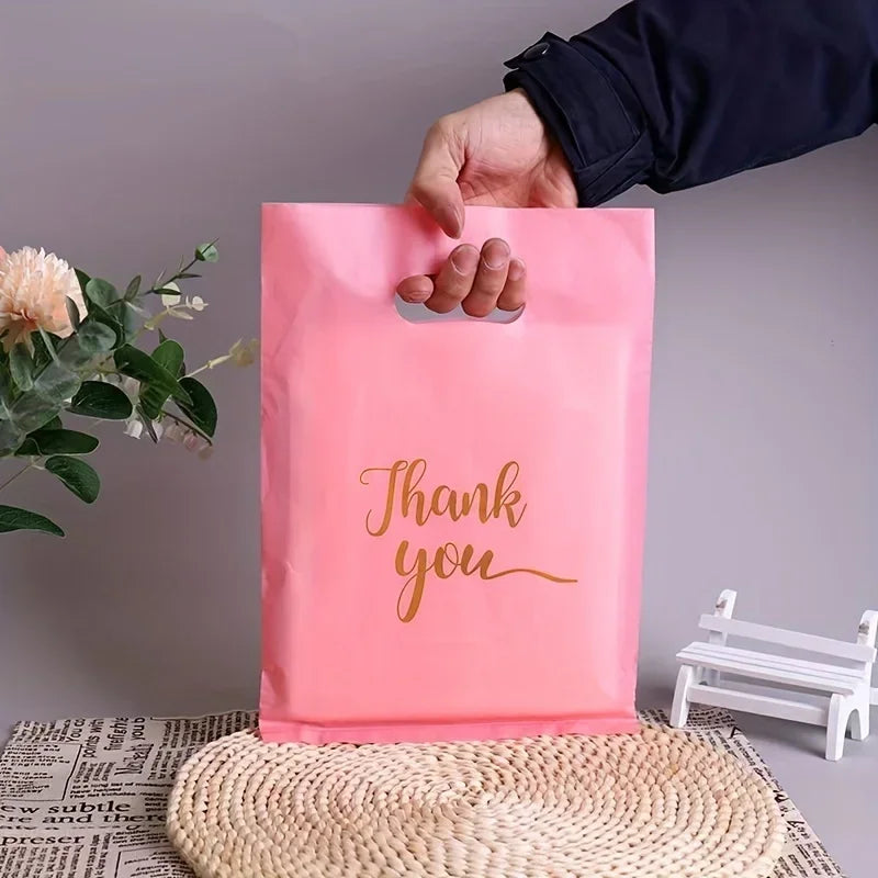 A hand is holding a pink clear plastic gift bag with gold lettering that says Thank you