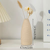 A minimalist beige ceramic vase holding dried wheat and a white daisy, displayed on a white shelf.