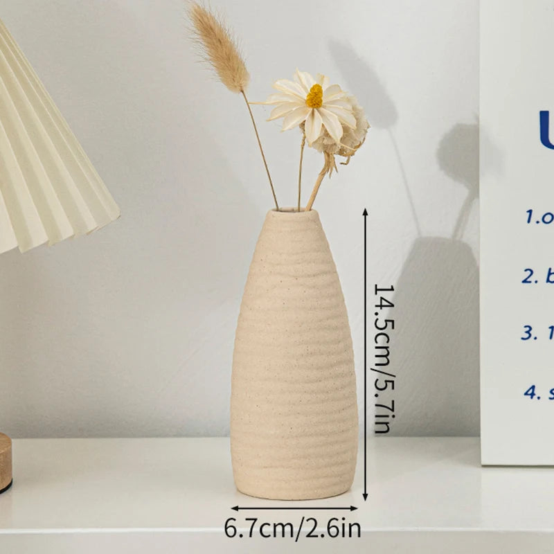 A minimalist beige ceramic vase holding dried wheat and a white daisy, displayed on a white shelf.