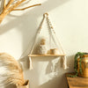 A macrame wood floating wall shelf with hanging rope displaying a small plant and candle on a light-colored wall.