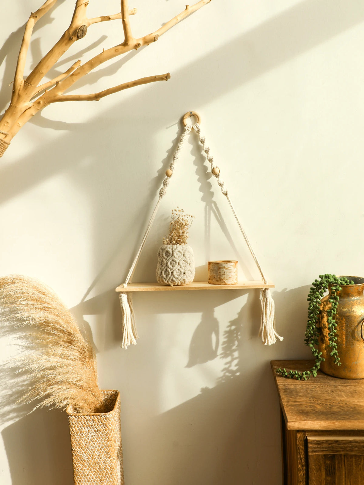 A macrame wood floating wall shelf with hanging rope displaying a small plant and candle on a light-colored wall.