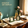 A vintage baroque resin picture frame rests on an open book and various gold jewelry on a wooden table.