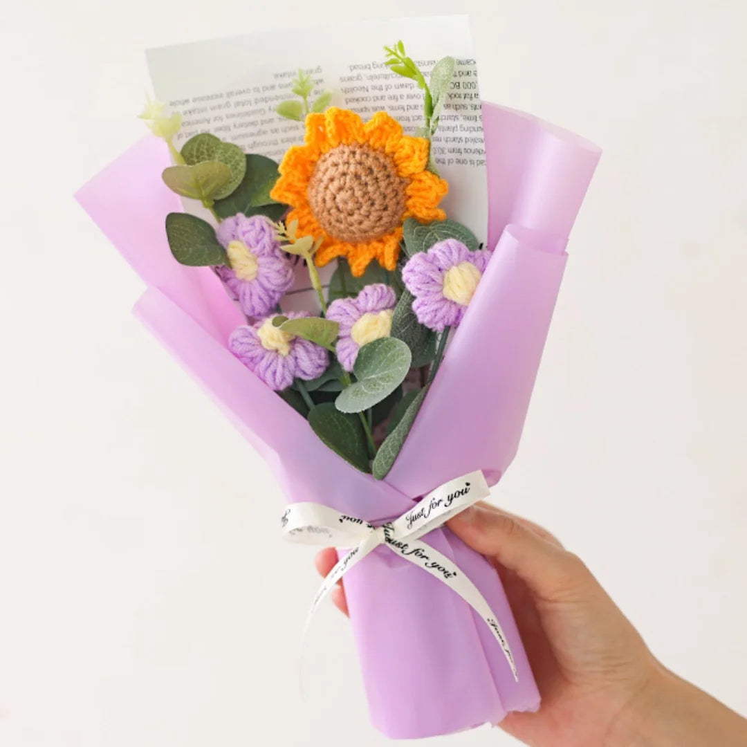 A hand holds a bouquet of handcrafted crochet sunflowers and purple flowers in a pink paper wrapping.