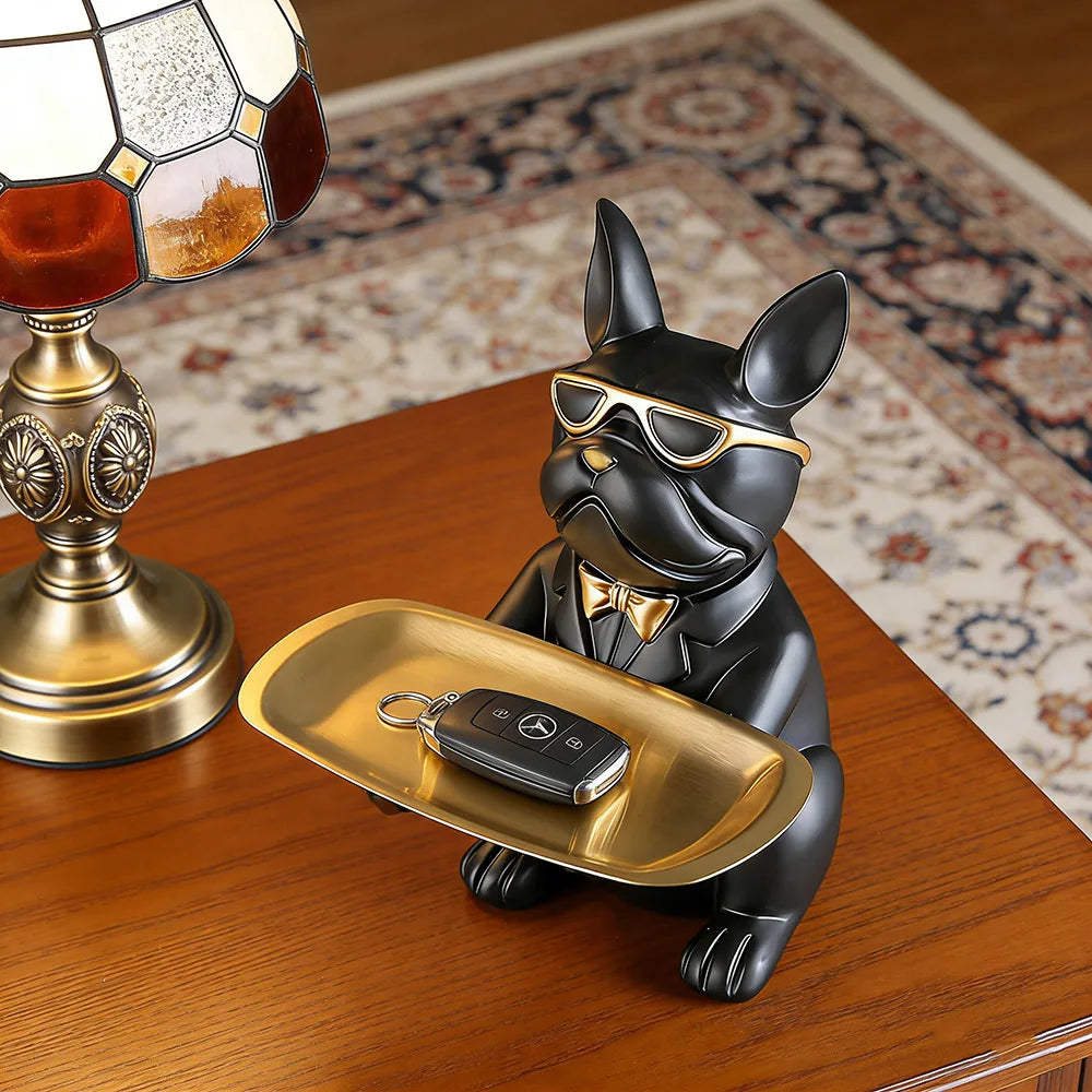 A black bulldog figurine wearing sunglasses is resting on a gold tray on a wooden surface.
