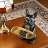 A black bulldog figurine wearing sunglasses is resting on a gold tray on a wooden surface.
