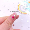 A hand holds a pink and white love heart flatback rhinestone mix for nail art, next to a rainbow-colored cutout.