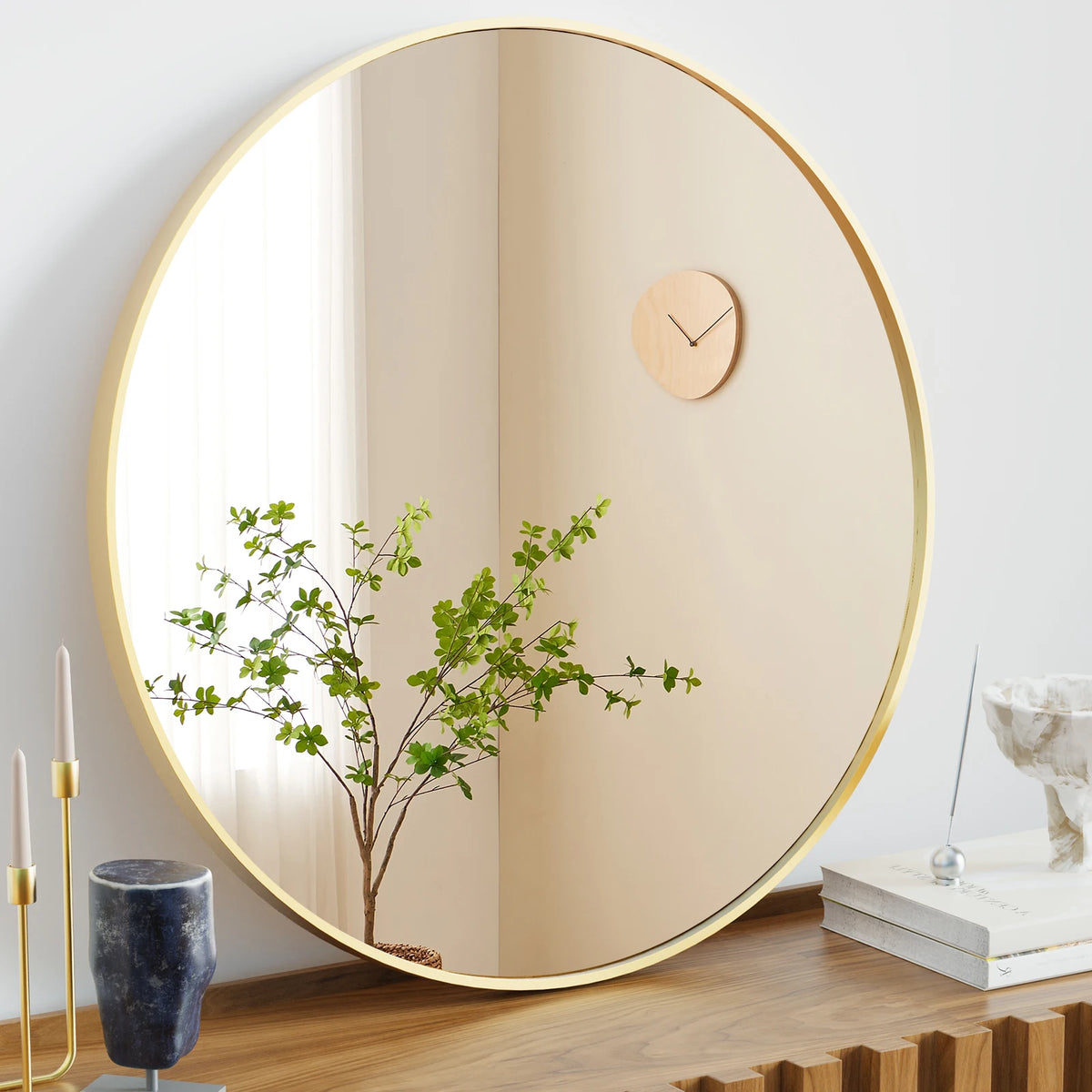 A large round vanity mirror with a gold-toned frame reflects a green plant and a wooden clock on a light surface.