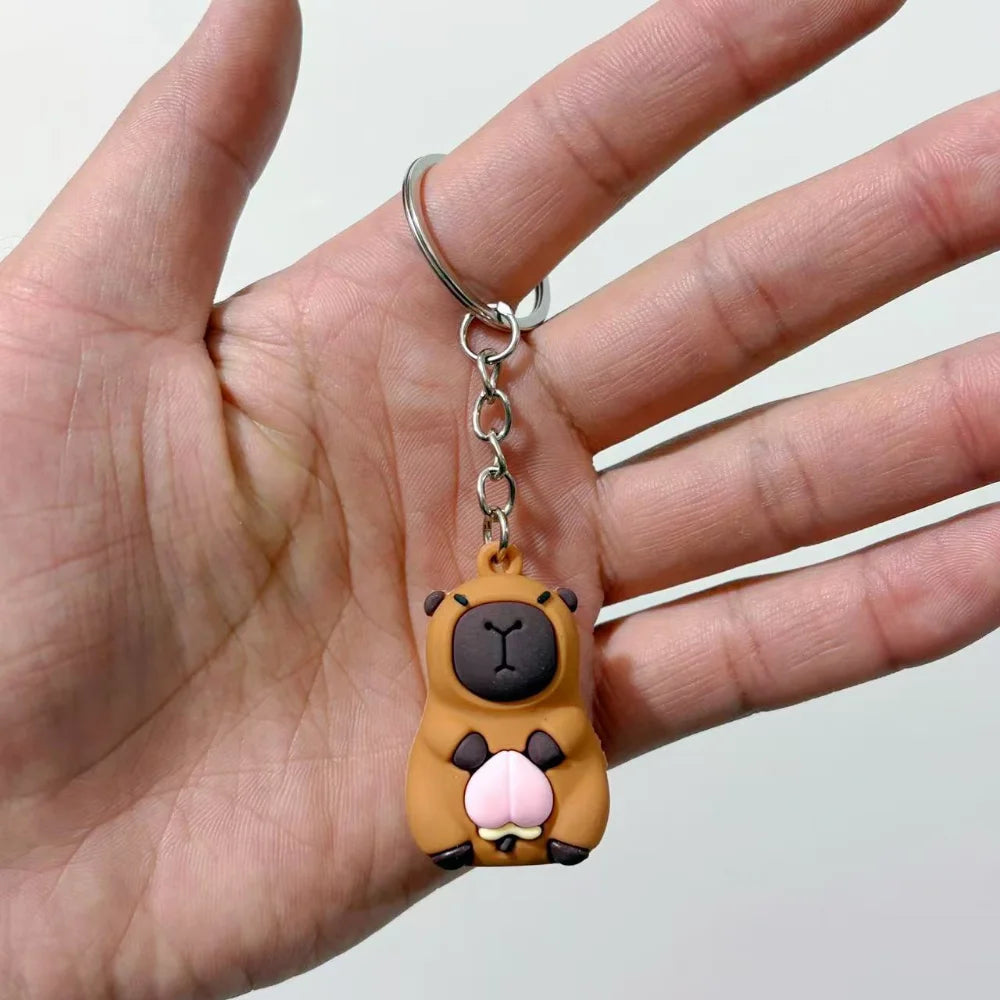 Un petit pendentif de clé en forme d'un capybara brun avec des accents roses, tenu par les doigts d'une personne.