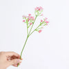 A hand holds a slender green stem with clusters of pink artificial baby's breath flowers.