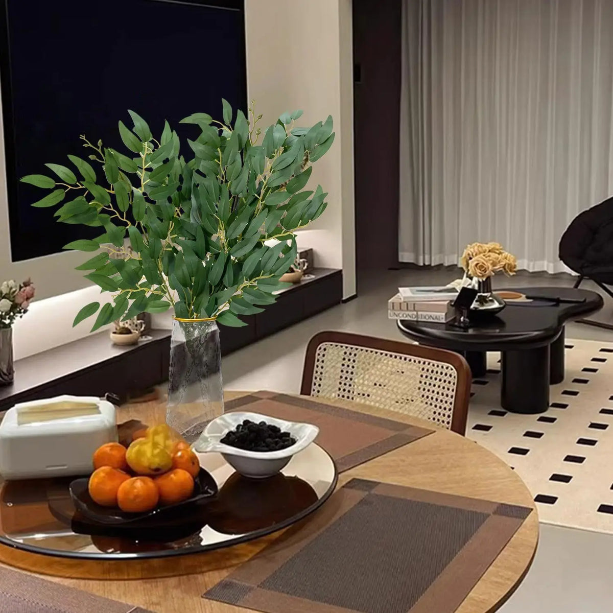Planta artificial de Ruscus italiano en maceta sobre una mesa con fruta y objetos decorativos.