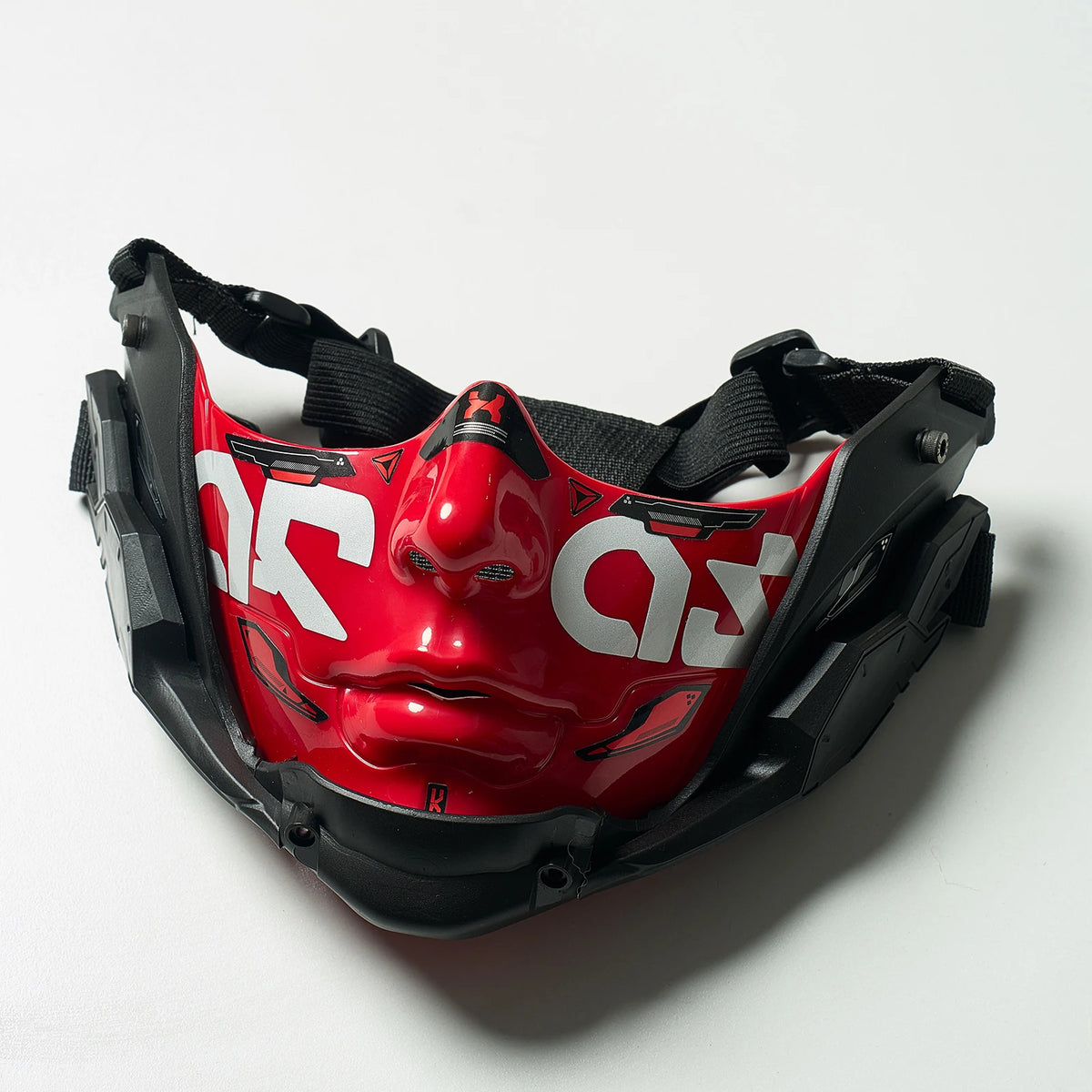 A red and black half face mask with a stylized face design is shown against a white background.