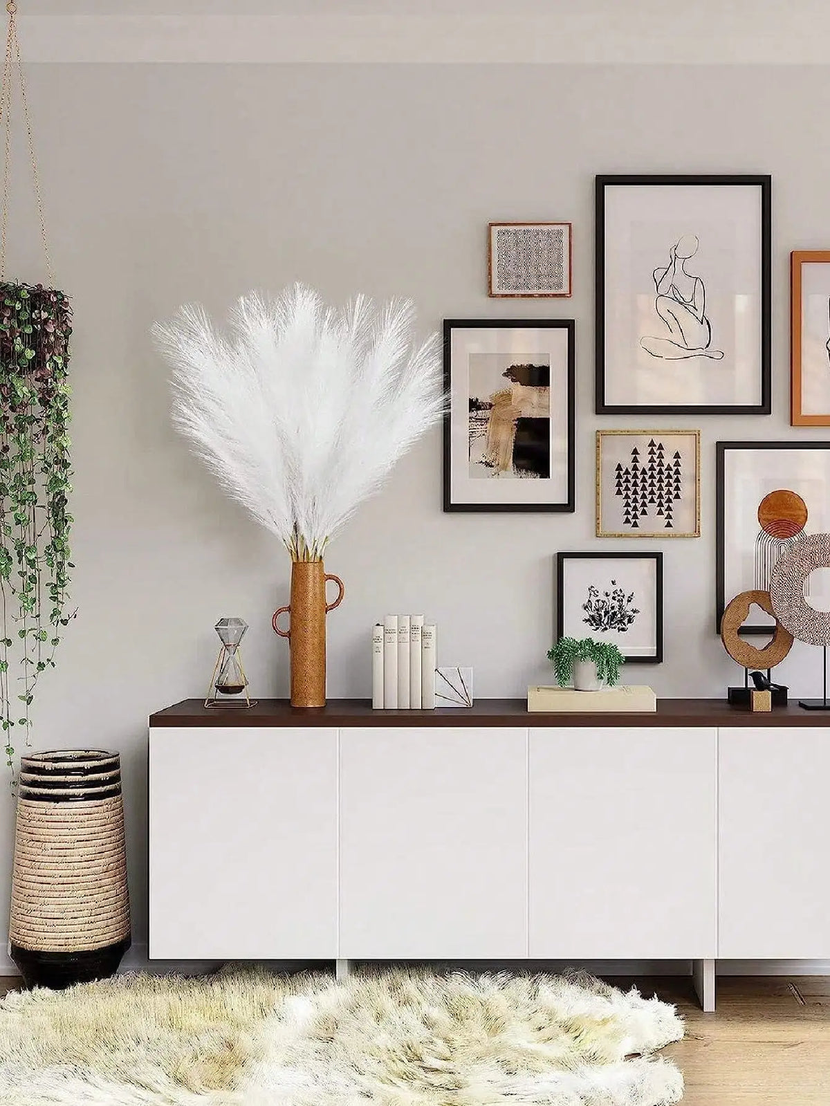 A display of white artificial reed stems in a gold vase against a light gray wall with framed artwork.