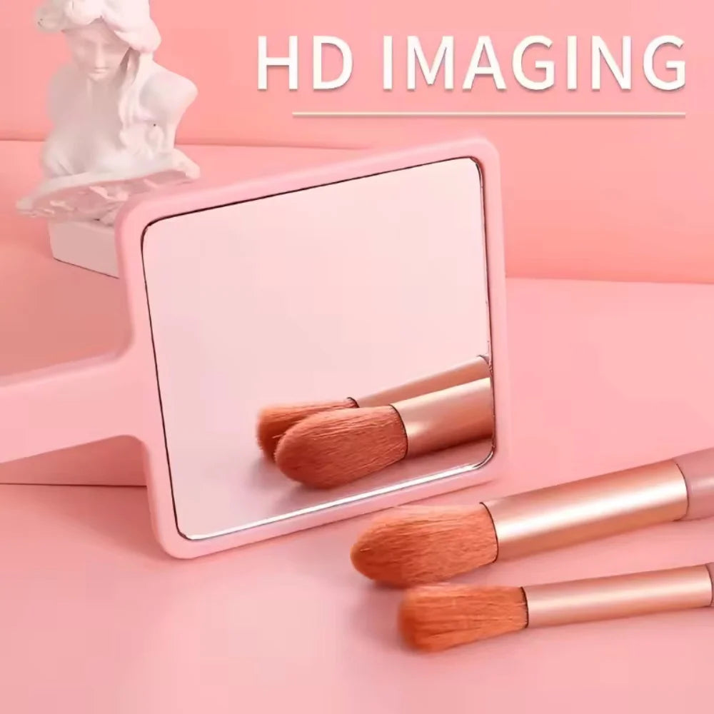 A pink compact handheld makeup mirror with a wooden handle and several makeup brushes is displayed on a pink surface.