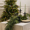 A long green artificial Norfolk pine garland draped on a white tablecloth with greenery and candles in the background.