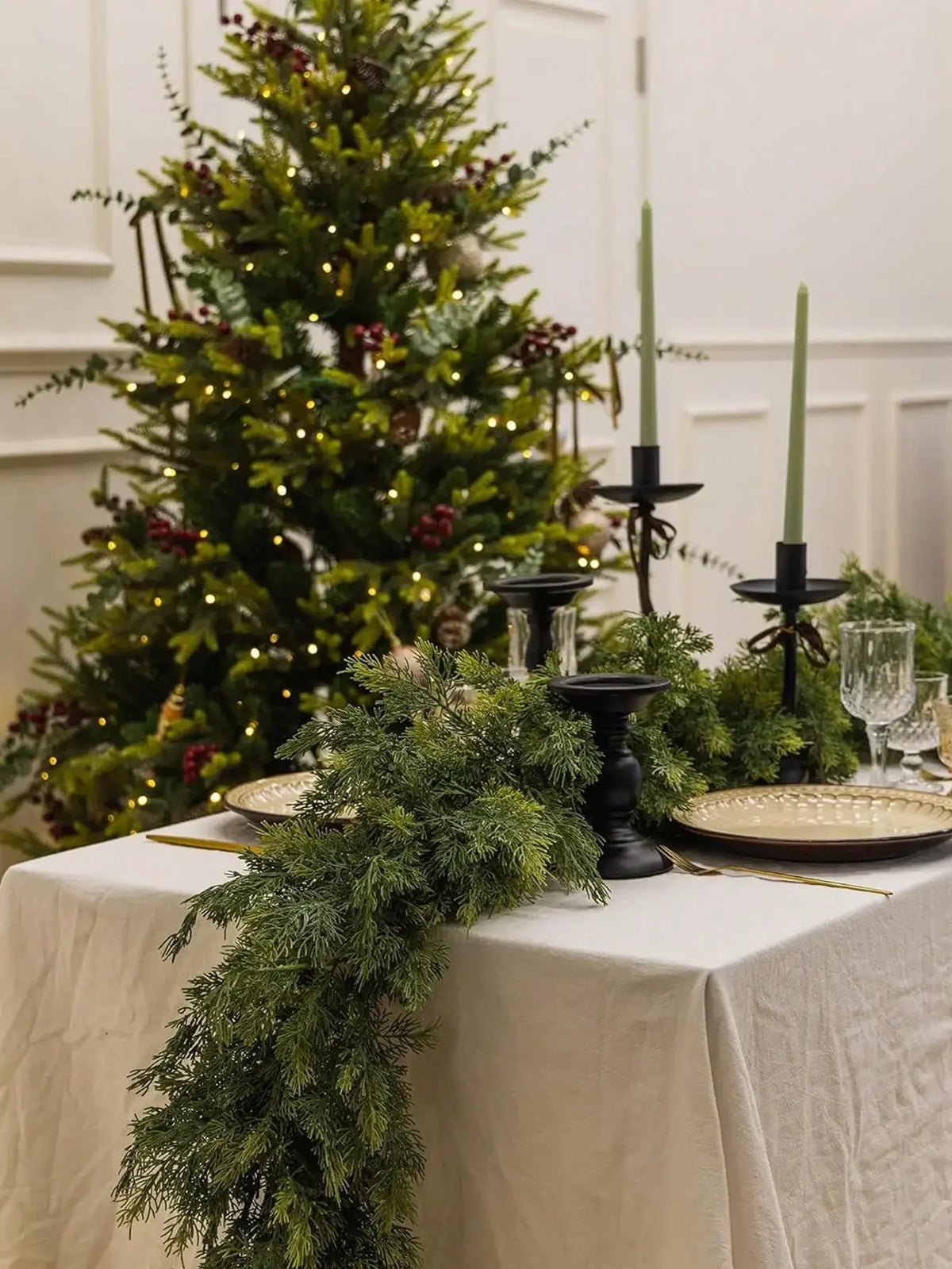 A long green artificial Norfolk pine garland draped on a white tablecloth with greenery and candles in the background.