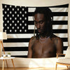 A young man with dreadlocks is shirtless, looking down, positioned in front of a black and white striped flag backdrop.