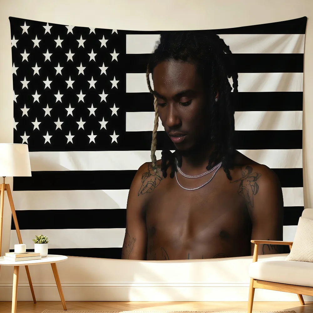 A young man with dreadlocks is shirtless, looking down, positioned in front of a black and white American flag backdrop.