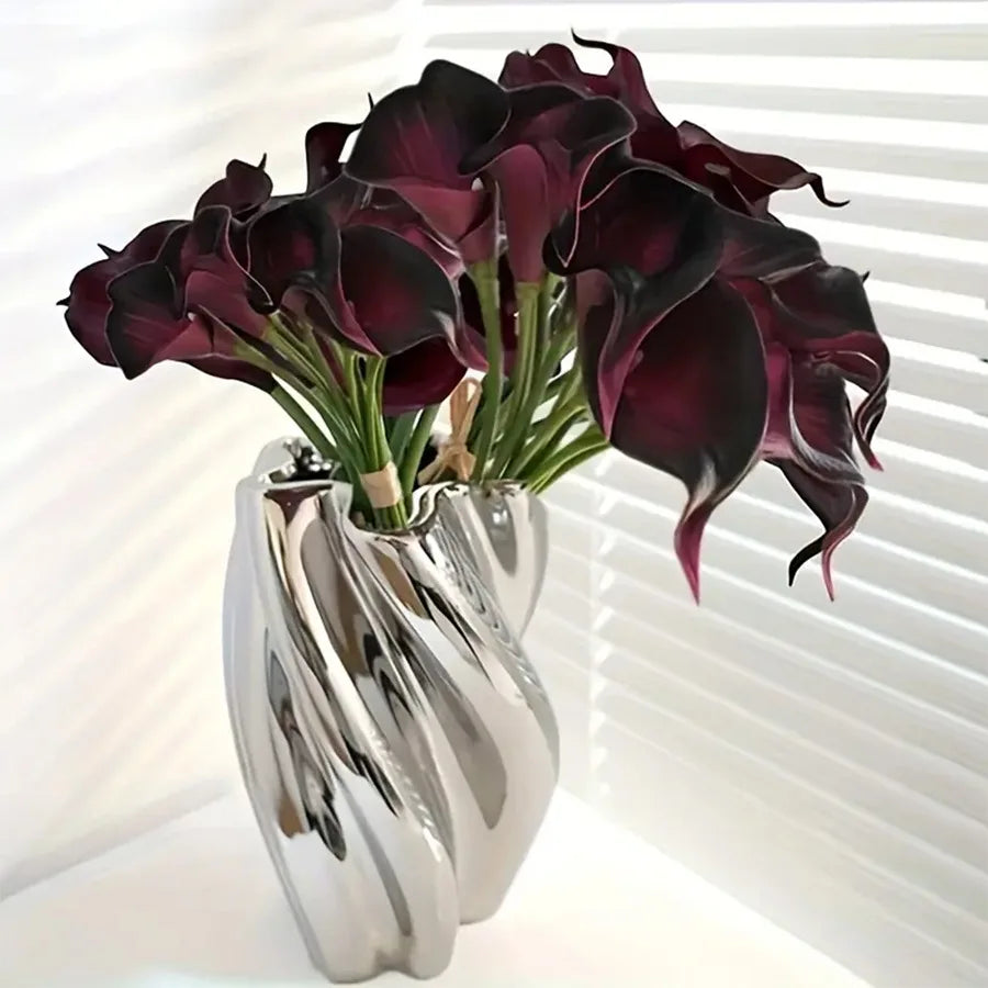 A bouquet of deep burgundy calla lily stems in a silver vase against a white background.