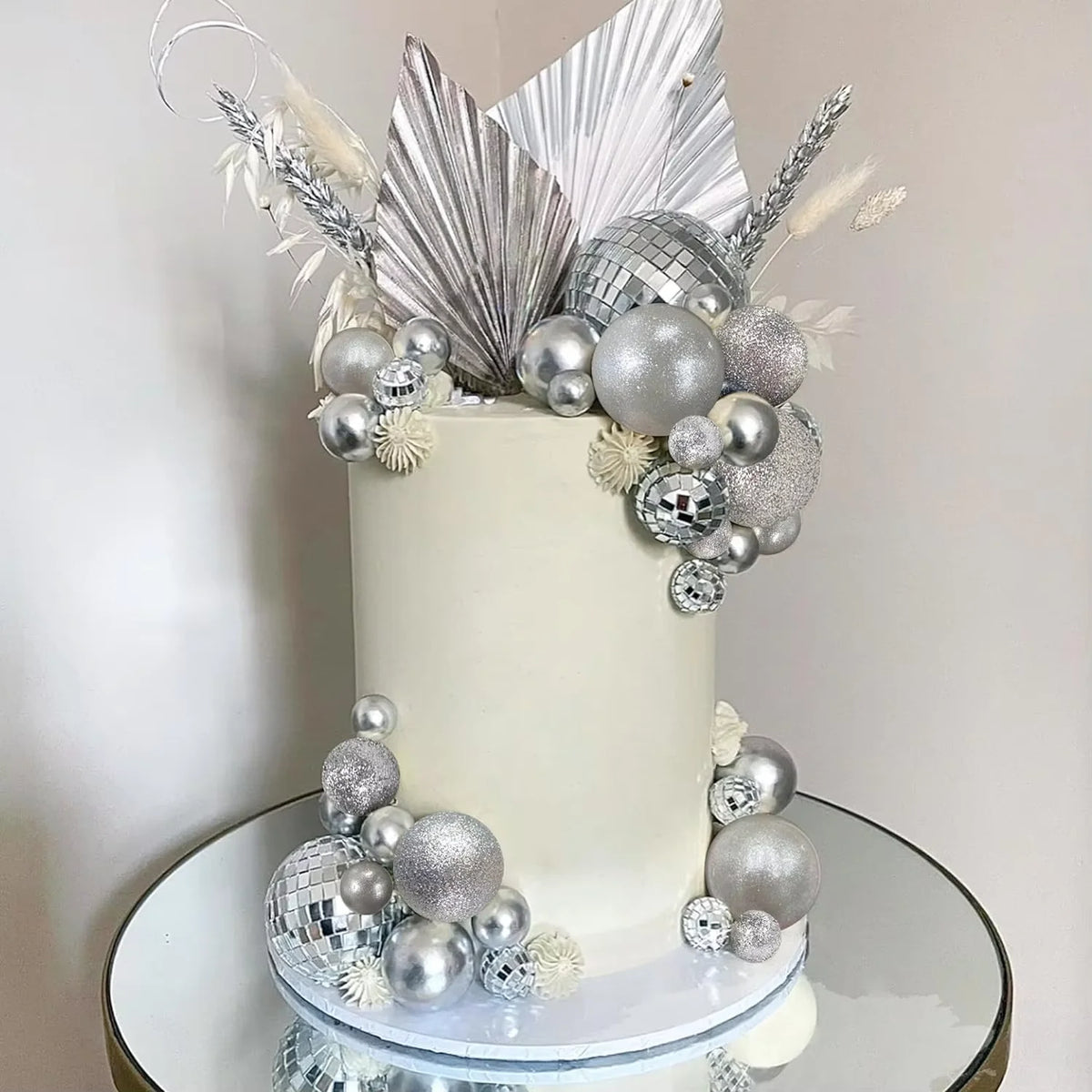 A white cake decorated with silver and gold disco ball-themed toppers and floral accents.