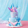 A tiered cake decorated with blue frosting, featuring mermaid tail toppers, seashells, and sea stars on a pink surface.