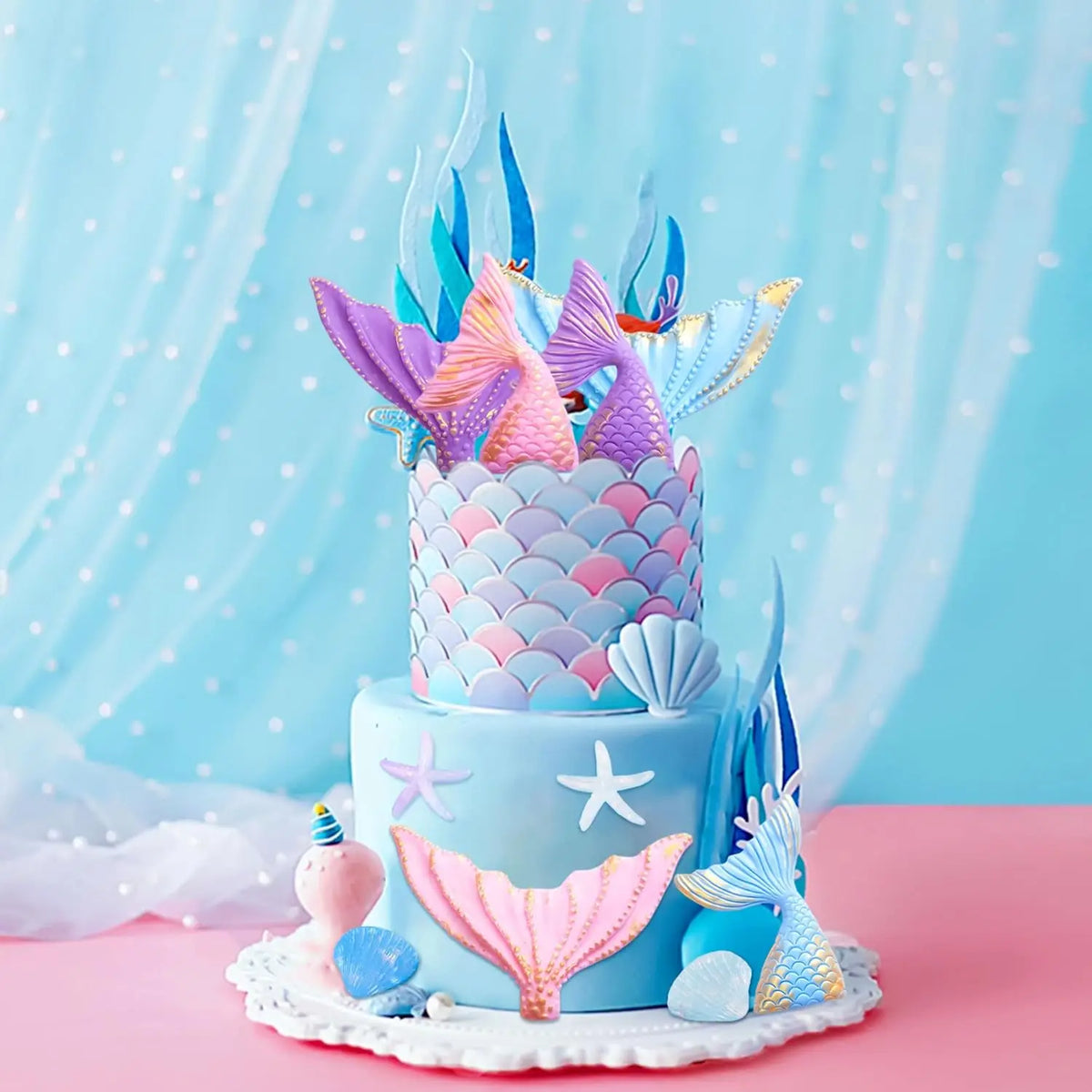 A tiered cake decorated with blue frosting, featuring mermaid tail toppers, seashells, and sea stars on a pink surface.