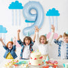 A large light blue foil number balloon shaped like 9 is displayed against a white wall with clouds and streamers.